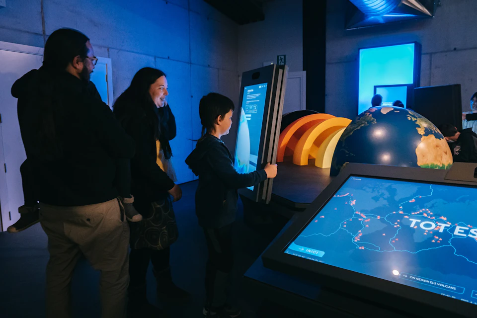 People interacting with interactive digital displays in a dark, blue-lit room, with colorful arches in the background.