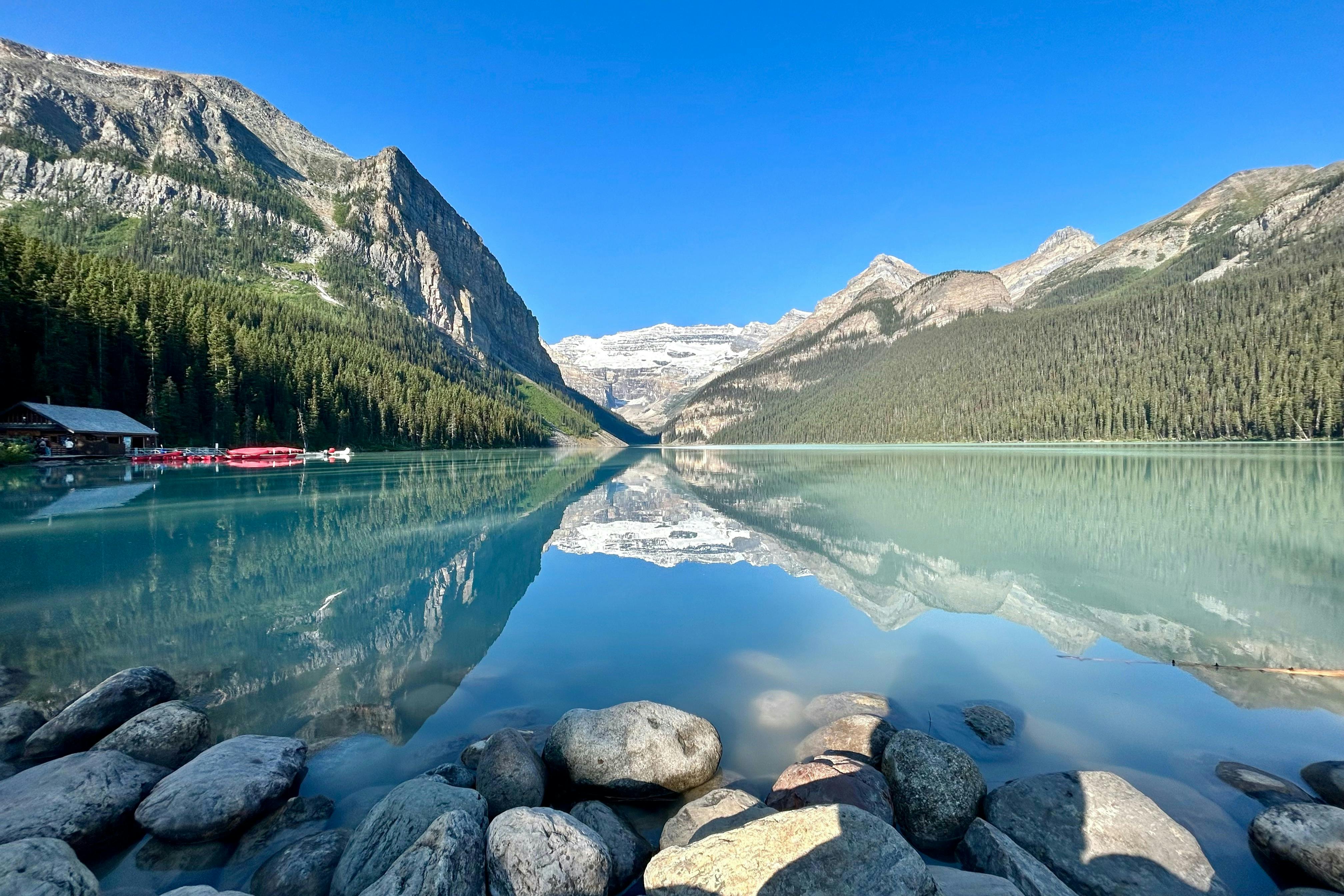 A tranquil lake with mountains and trees reflected in the clear water, rocks in the foreground, and a dock with boats on the left.