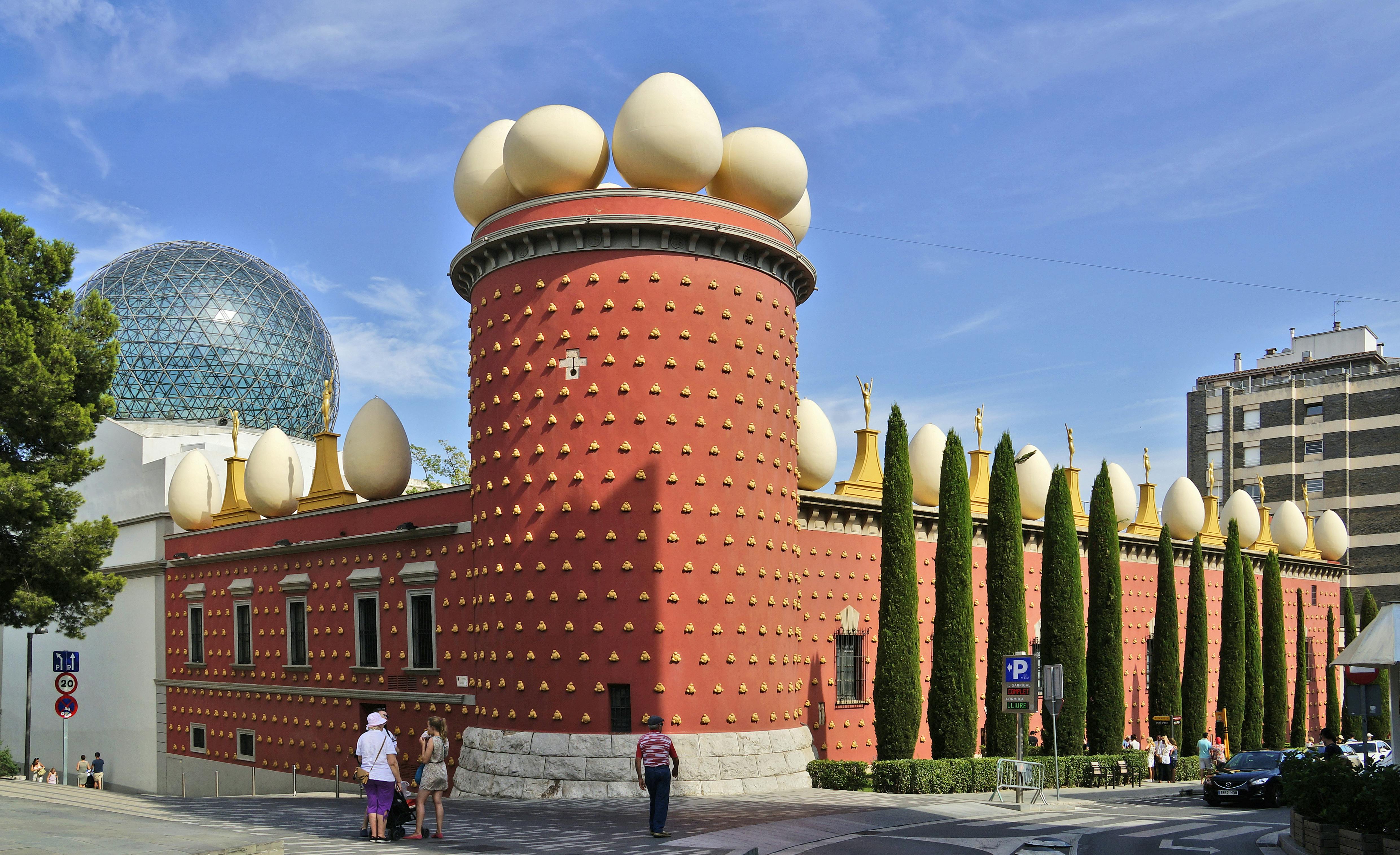 Red building adorned with large white egg sculptures and golden statues, surrounded by tall trees; people strolling nearby.
