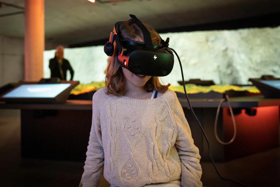 A child wearing a virtual reality headset and a beige sweater, standing in a dimly lit room with blurred background.