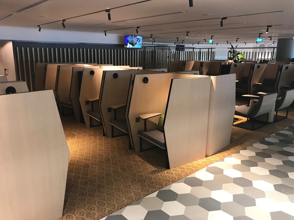 Modern lounge area with rows of light wood privacy booths, hexagonal floor tiles, and a TV mounted on the back wall.