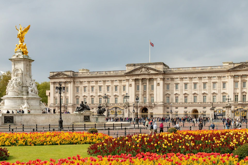 A large historic building with a flag on top, a fountain, and colorful flowerbeds in the foreground under a cloudy sky.