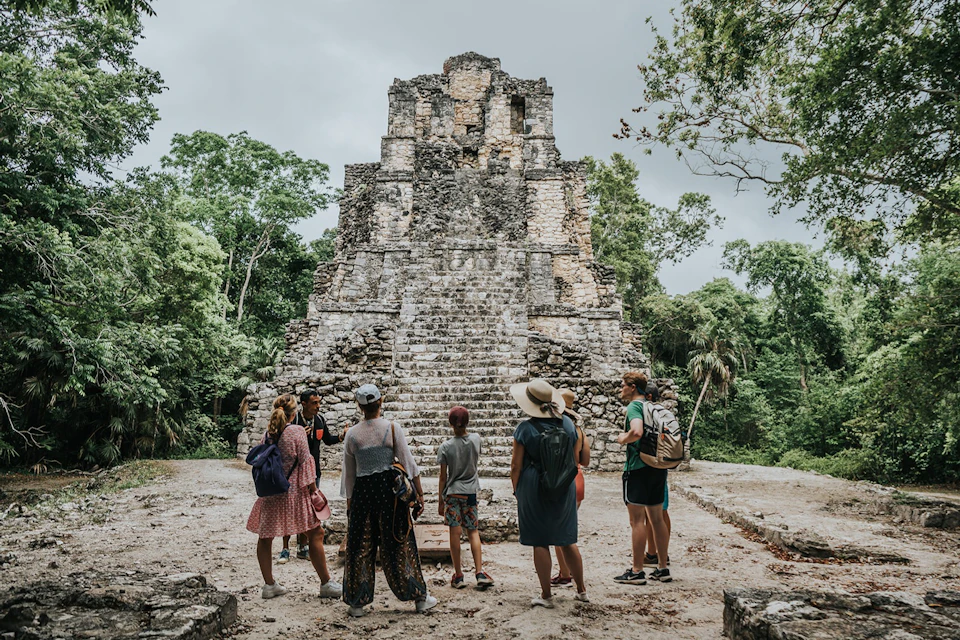 A group of people stand near an ancient stone pyramid surrounded by lush greenery on a cloudy day.