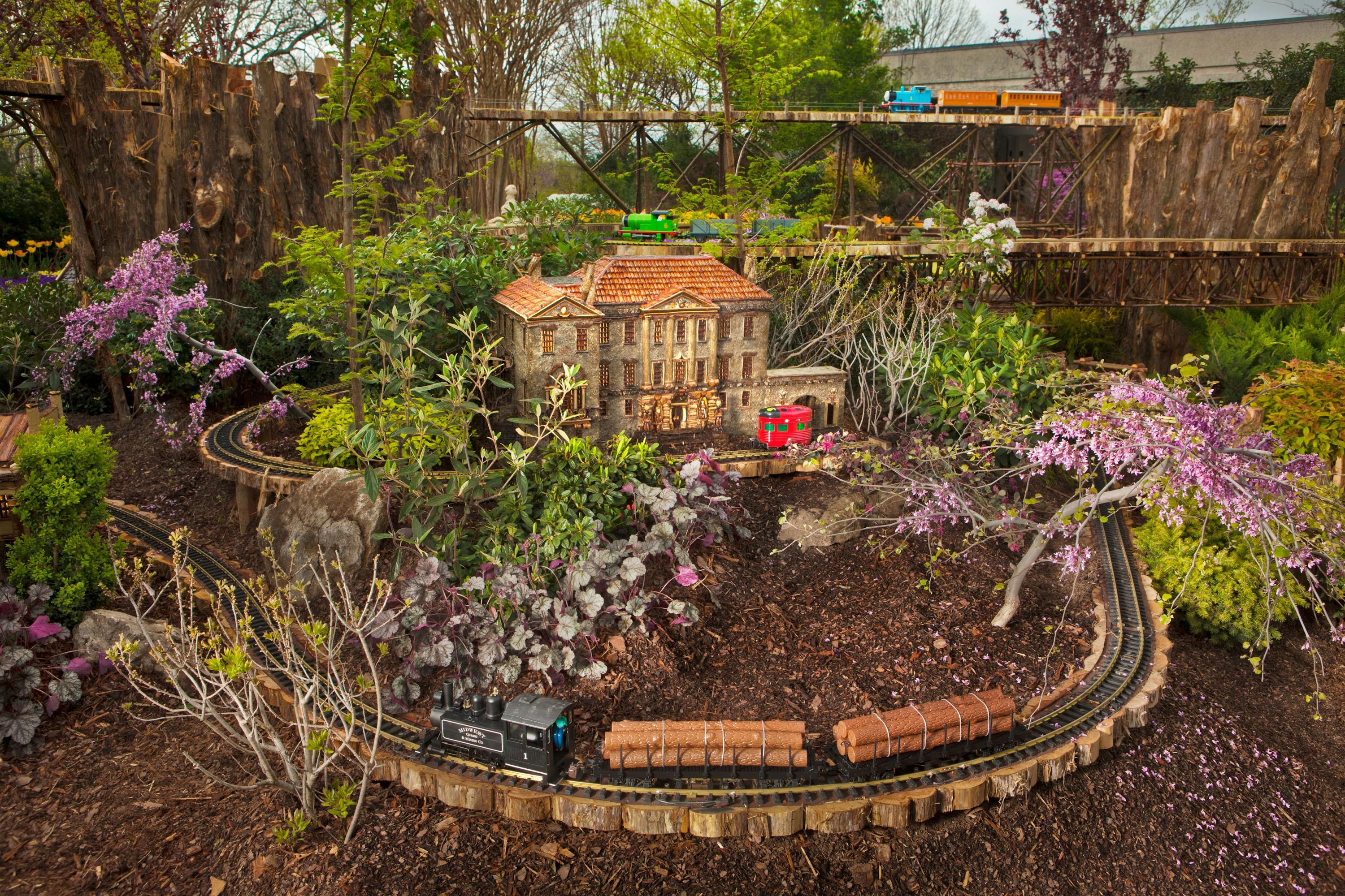 Zahradní miniaturní scéna s malou modelovou budovou, vlaky na kolejích a detailní vegetací a terénními úpravami.