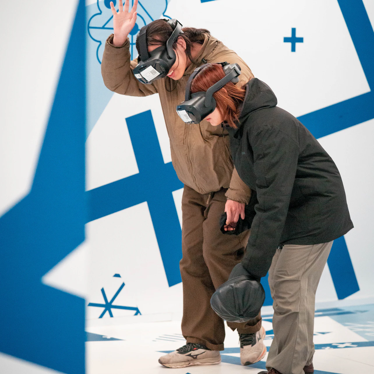 Two people wearing virtual reality headsets interact in a room with blue and white geometric patterns.