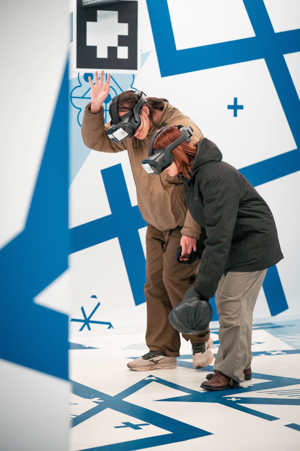 Two people wearing virtual reality headsets interact in a room with blue and white geometric patterns.