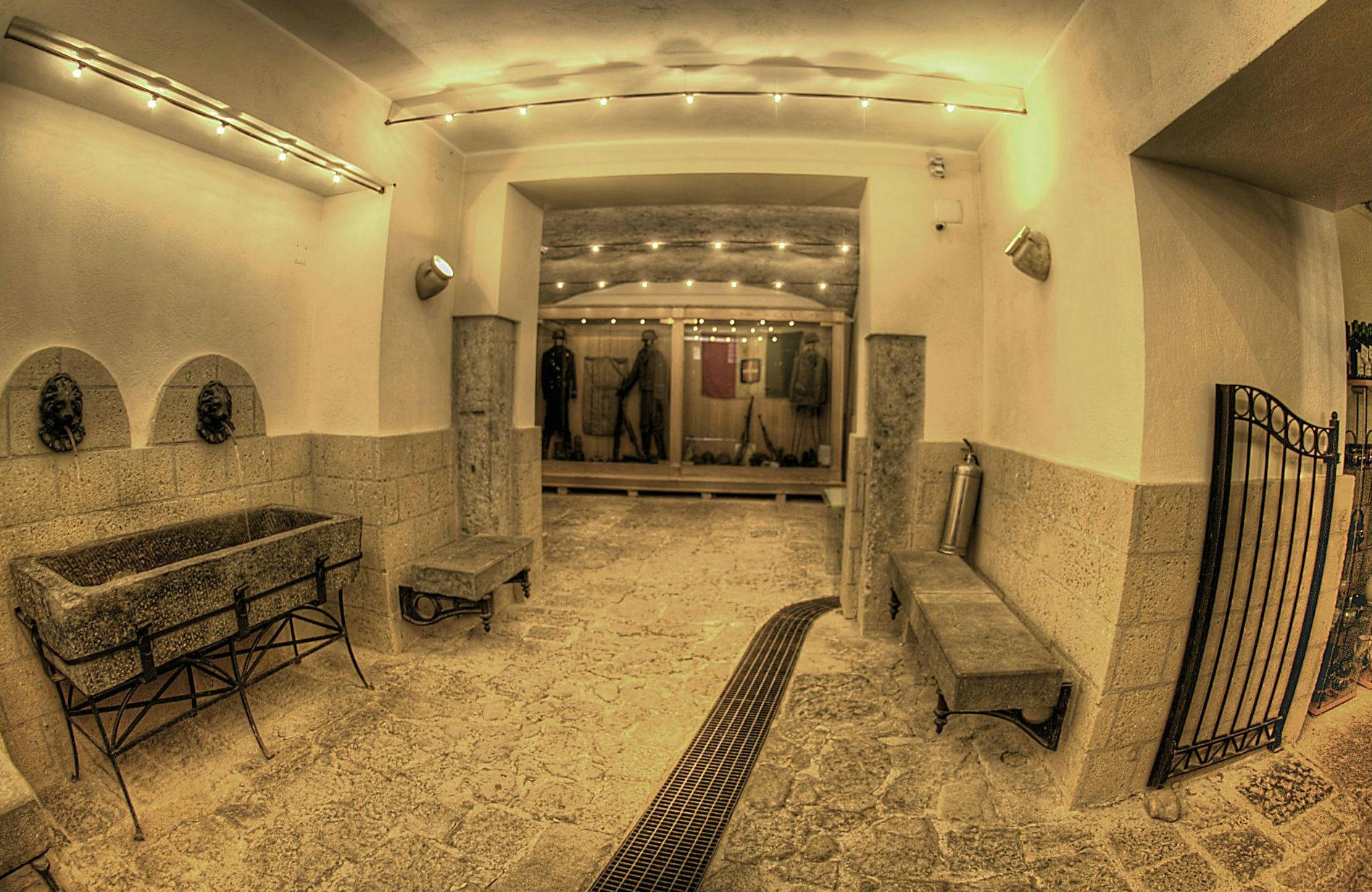 Salle aux murs de pierre avec des uniformes militaires historiques sur des mannequins. Des bancs, un bassin et un éclairage tamisé créent une ambiance de musée.