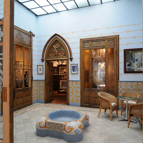 Ett dekorativt atrium med kaklade väggar och golv, en mosaikfontän, korgstolar och trädörrar med glaspaneler.