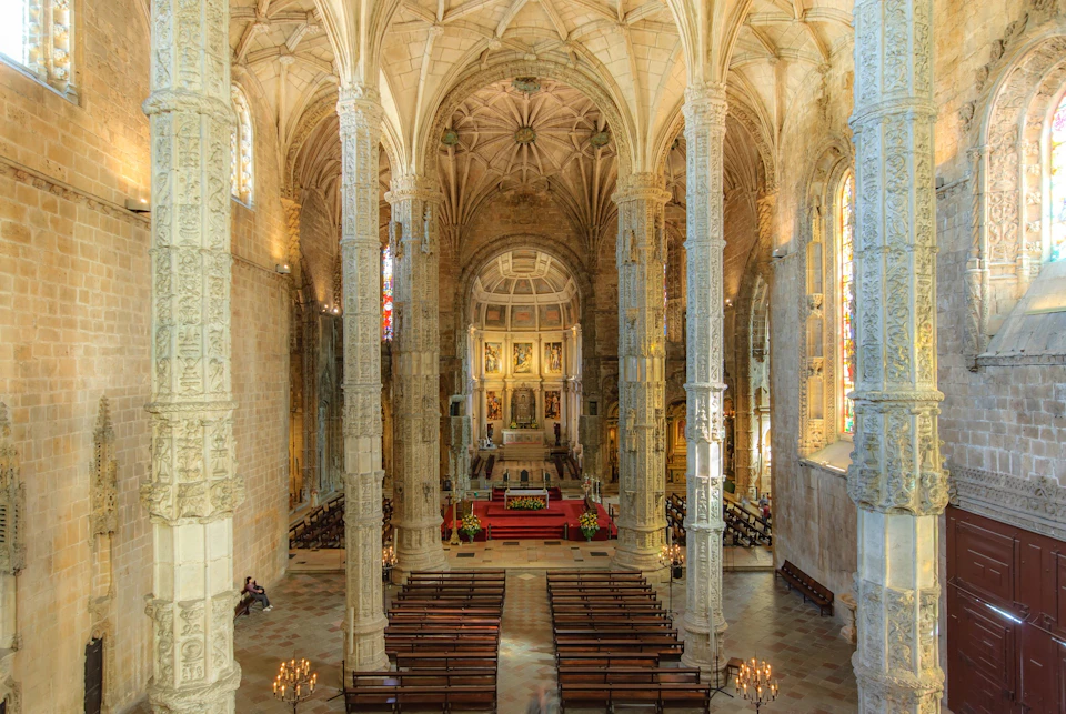 Interior of a grand cathedral with ornate columns, vaulted ceiling, wooden pews, and an altar adorned with paintings and flowers.