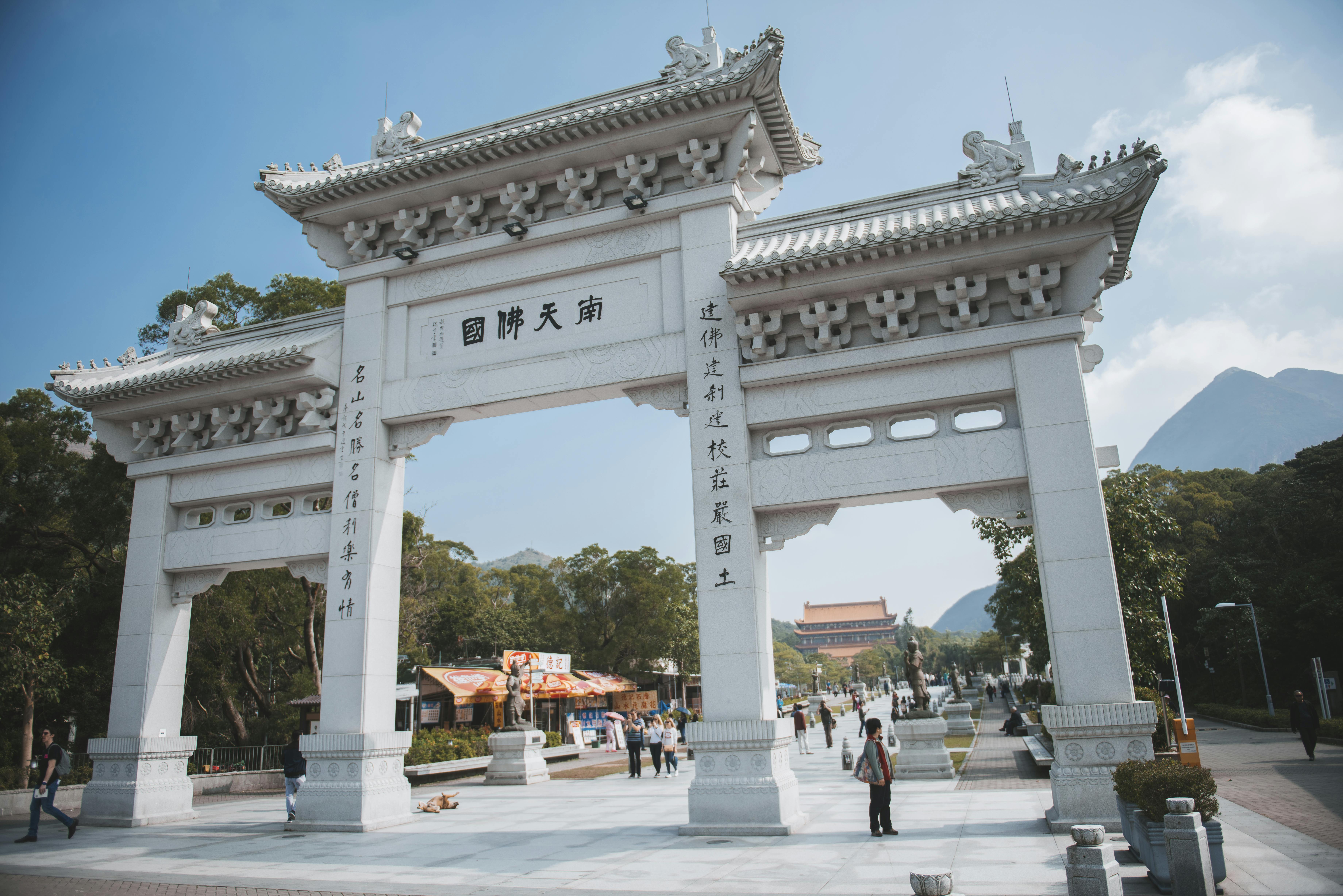 Large ornate archway with Chinese inscriptions, people walking below, trees, and buildings in the background, under a clear sky.