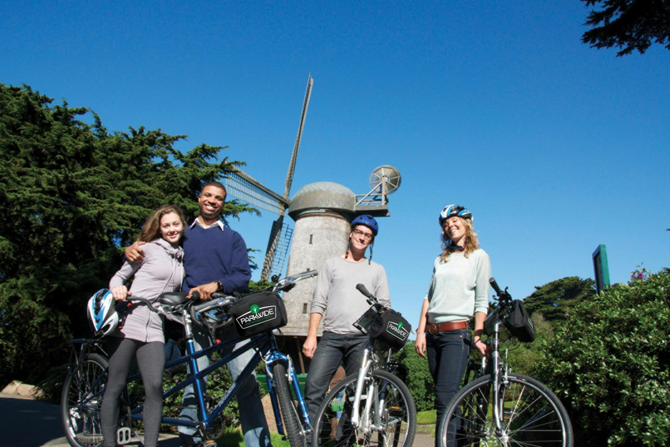 Four people with bicycles standing in front of a windmill, two of them wearing helmets. Trees and blue sky in the background.