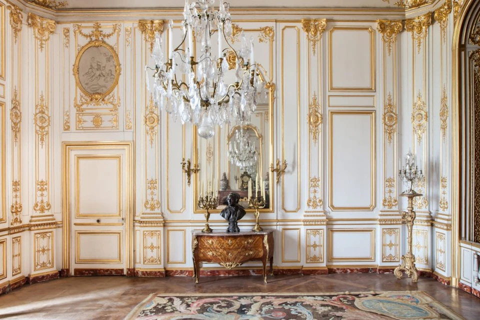 Ornate room with gold detailing, a large crystal chandelier, a mirrored wall, and a wooden table with a bust and candlesticks.