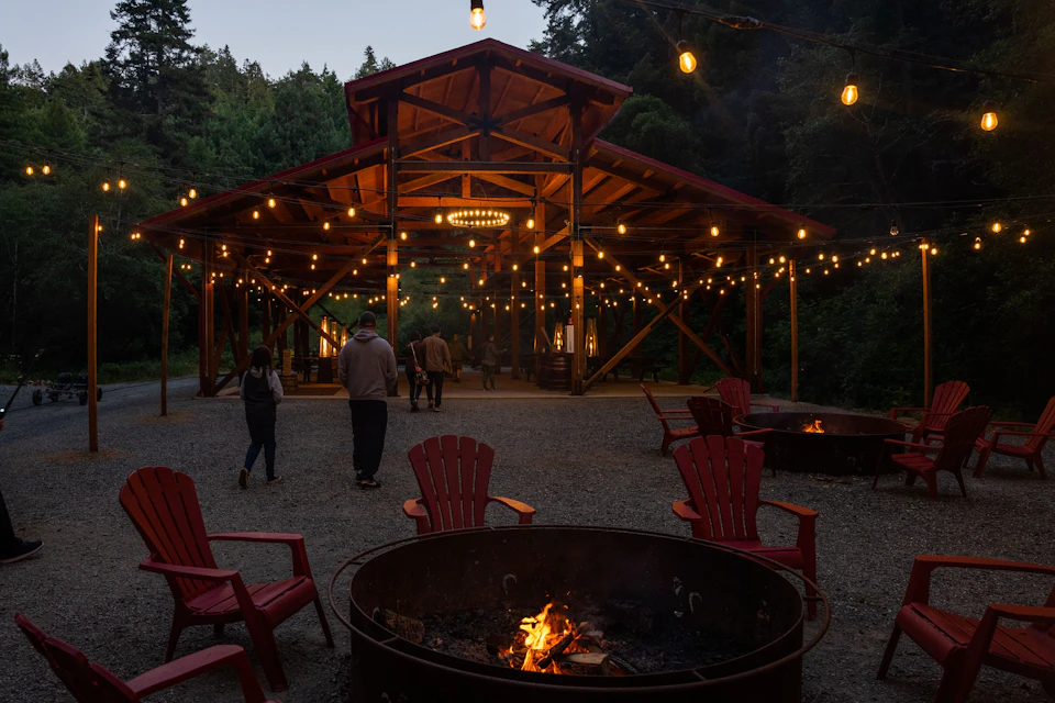 People walk towards a pavilion with hanging lights, surrounded by red chairs and fire pits under a twilight sky.