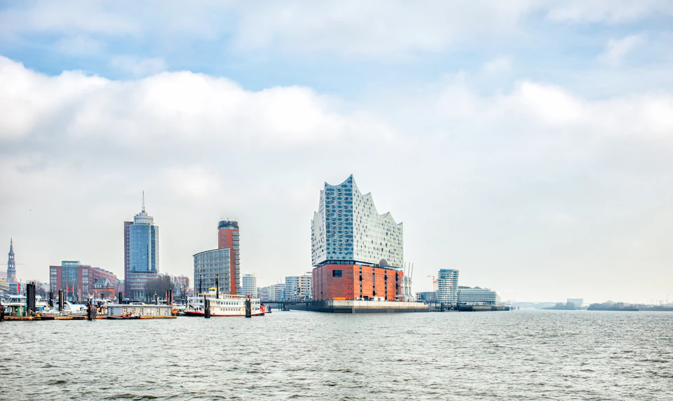 Modern waterfront buildings with unique architecture, including a wavy-roofed structure, viewed across a waterway under a partly cloudy sky.