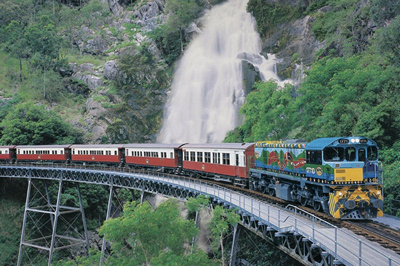 一列带有红色和白色车厢的火车在桥上行驶，后面是郁郁葱葱的绿地上的瀑布。