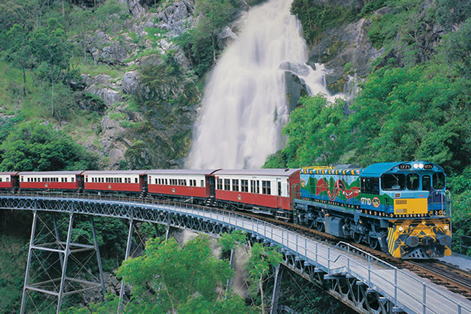A train with red and white carriages travels on a bridge with a waterfall in the background amidst lush greenery.