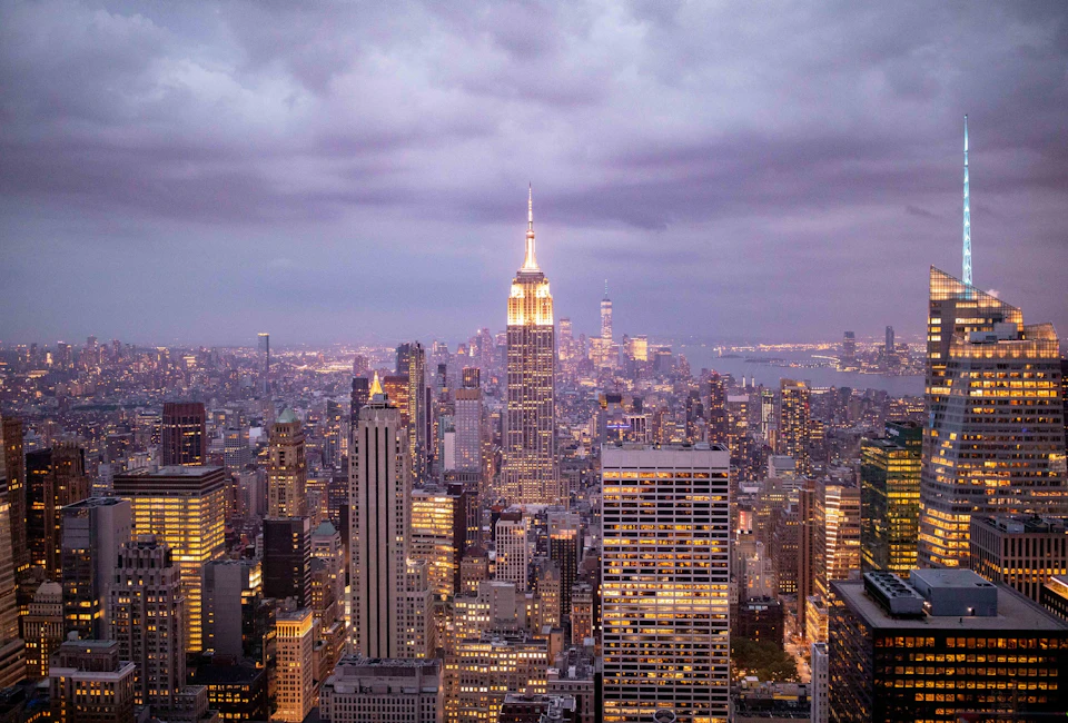 Cityscape view of New York City at dusk with the Empire State Building illuminated against a cloudy sky.