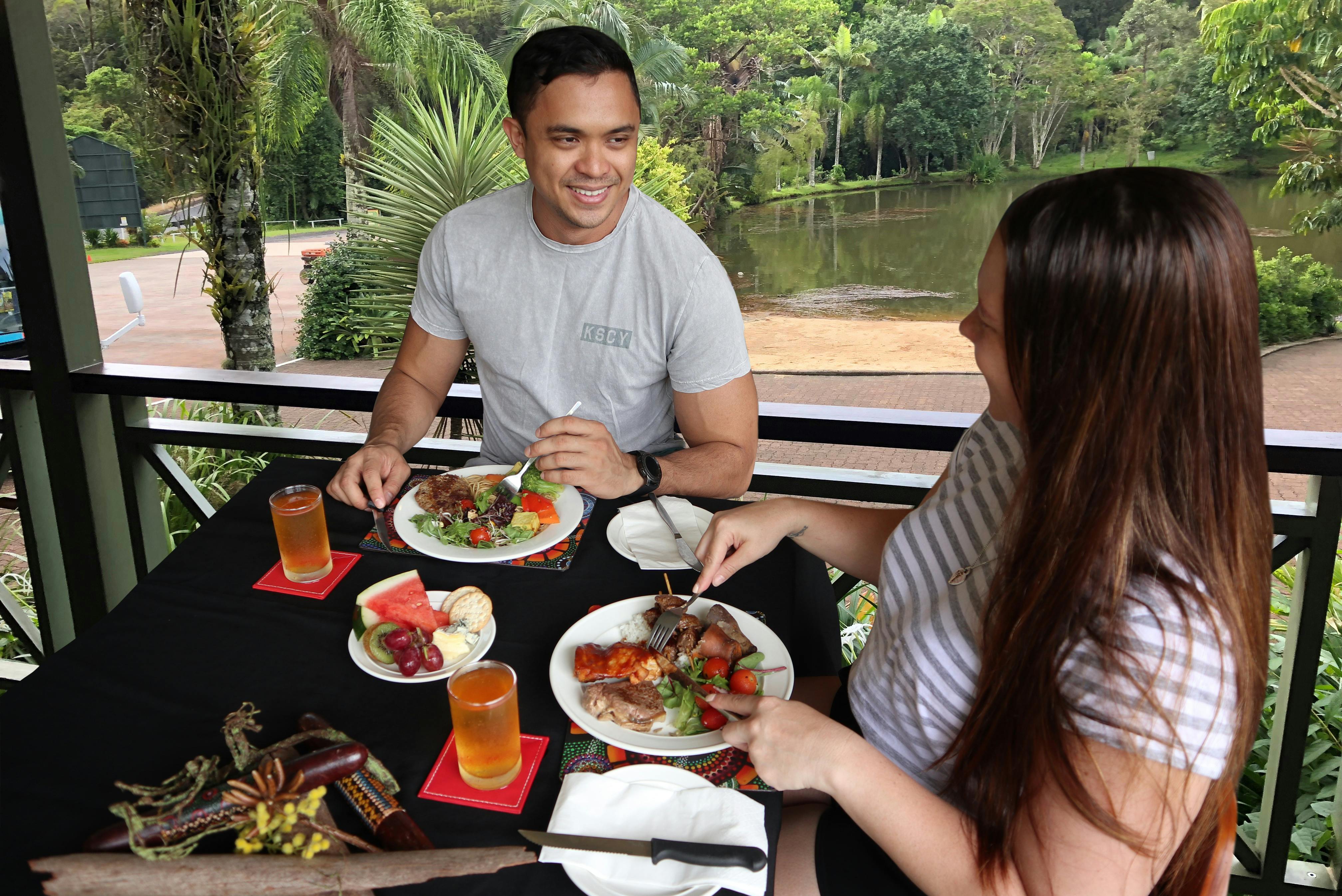 两人在户外餐桌上享用美食、饮料和水果。 背景中有郁郁葱葱的绿地和池塘。