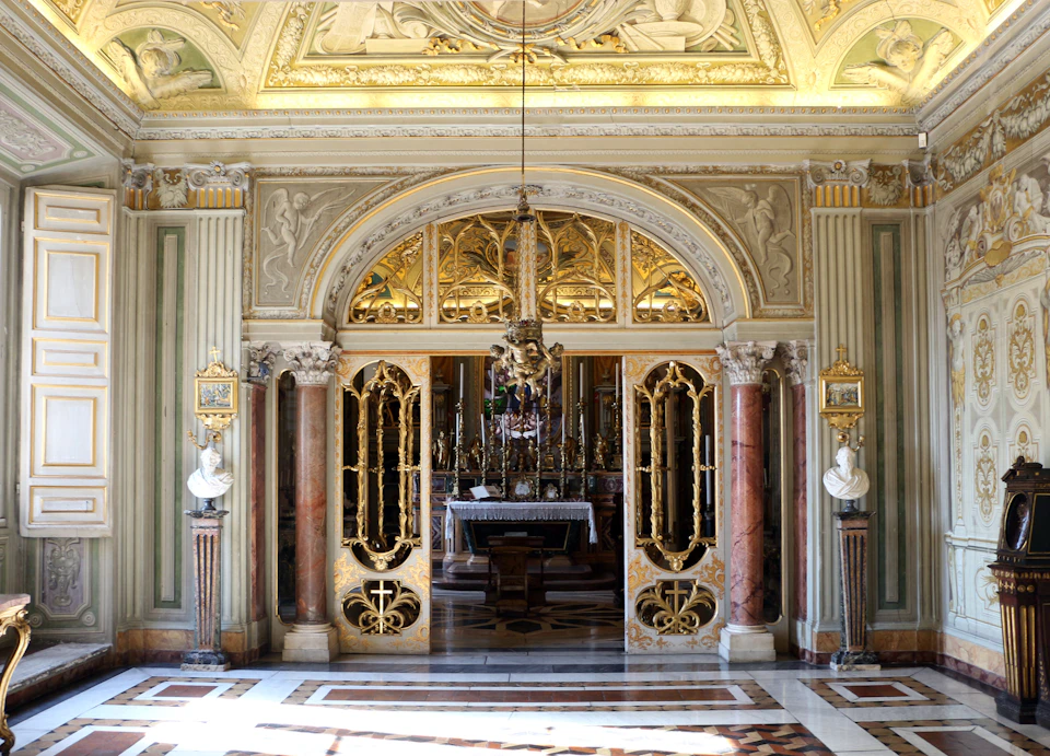 Ornate, gilded chapel doors framed by marble columns within a richly decorated room featuring sculptures and intricate ceiling details.
