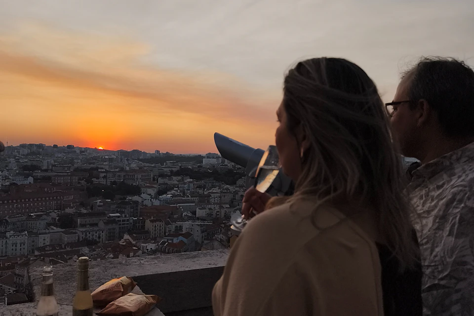 A person with long hair holds a glass, watching a cityscape at sunset from a rooftop with a telescope nearby.