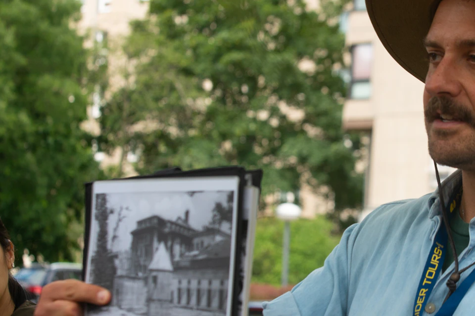 A person holds up a black-and-white photo while speaking to a small group outdoors in an urban area with trees and buildings.