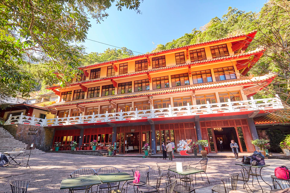 A large red and yellow multi-story building with balconies, surrounded by greenery. Outdoor seating and people are visible.