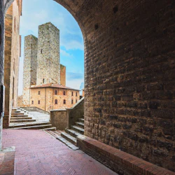 En stenbelagd gångväg med trappsteg som leder till höga medeltida torn mot en blå himmel, sedd genom en tegelvalvgång.