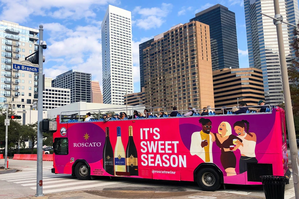 A double-decker bus with vibrant Roscato wine advertisements passes by skyscrapers under a partly cloudy sky.