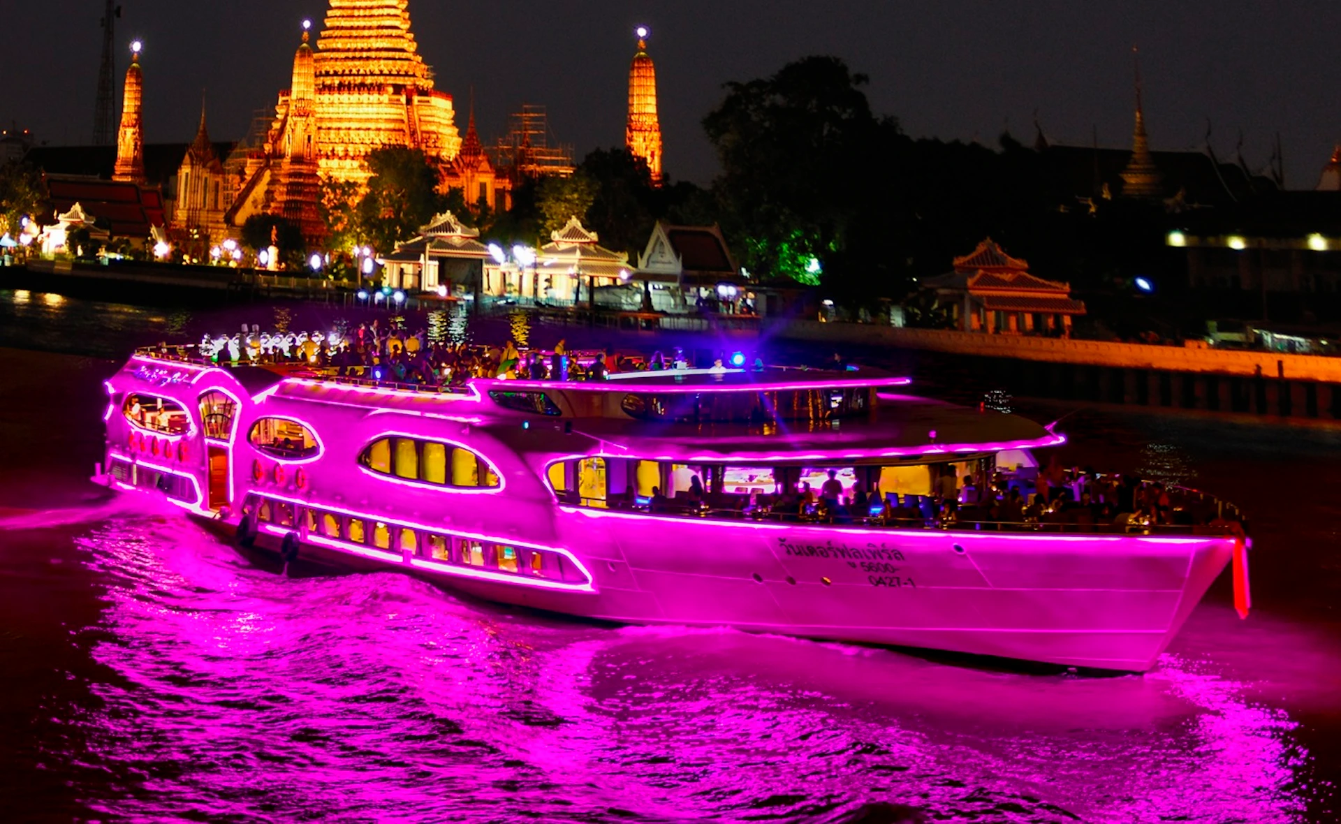 A riverboat illuminated with pink neon lights sails at night near a brightly lit temple complex.