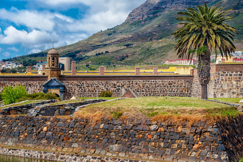 A stone fort with a grassy top, a bell tower, and a palm tree, with a mountain and cloudy sky in the background.