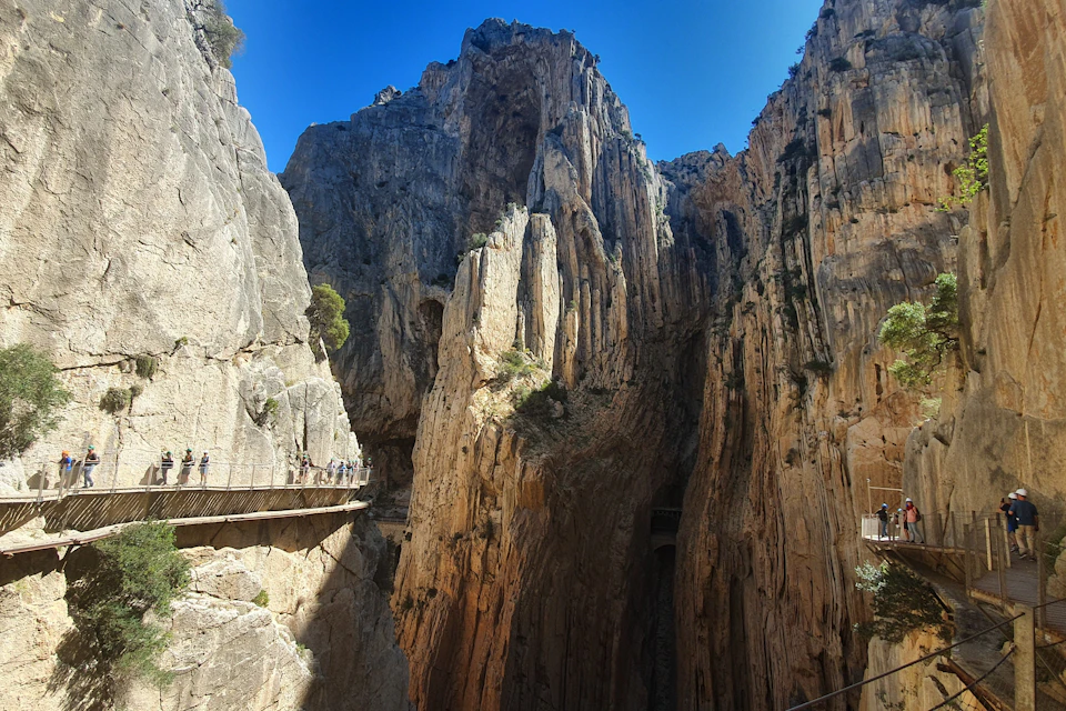 A narrow suspended walkway along a steep rocky canyon with towering cliffs under a clear blue sky.