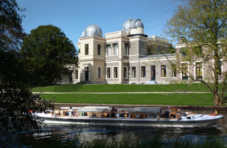 A boat with passengers sails on a calm river in front of a historic building with two domed observatories under a clear blue sky.