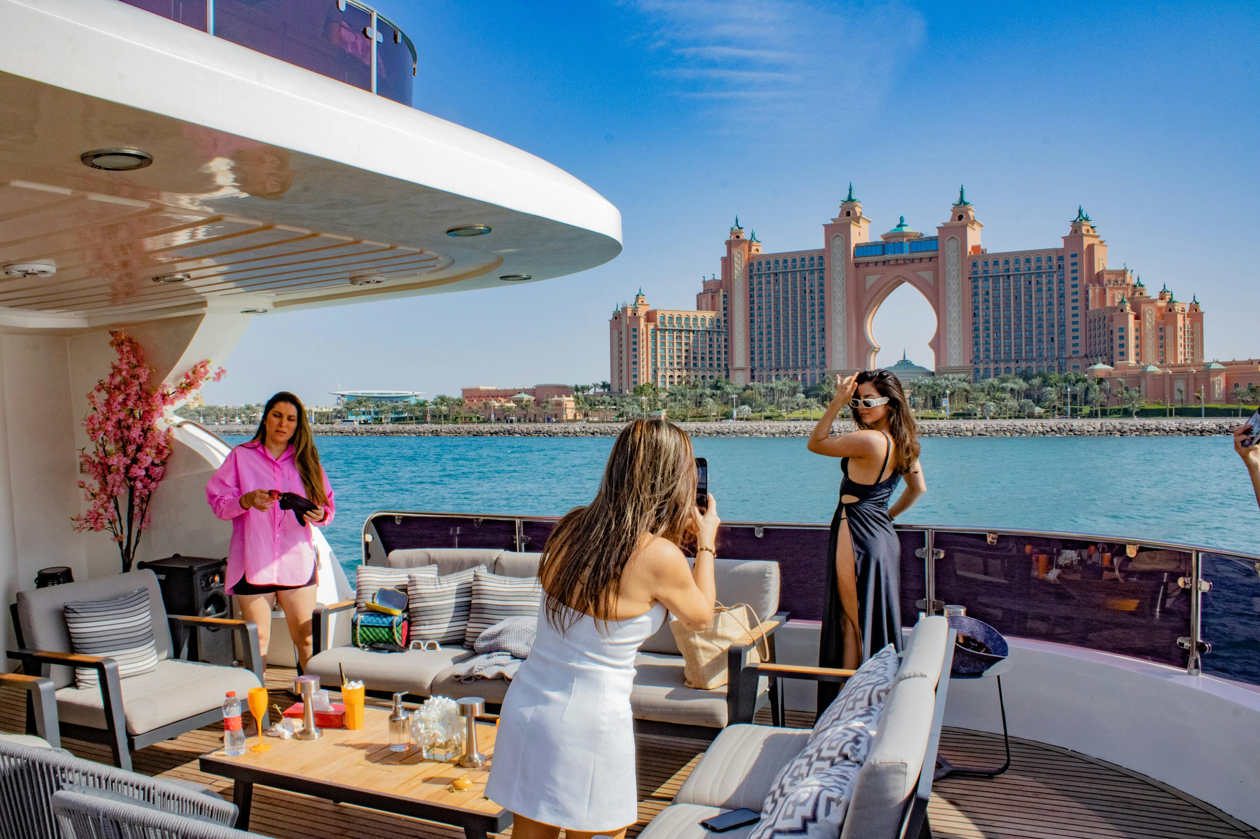 Two women on a luxury yacht; one in pink, the other taking a photo. Background includes an ocean view and a tall, pink building.