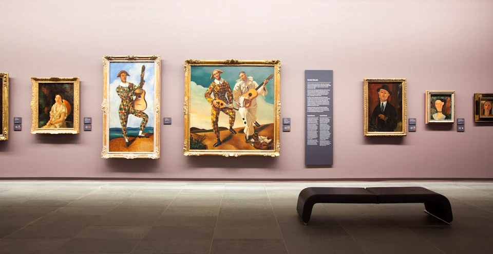 Three framed paintings on a museum wall, one with label text and a bench in the foreground.