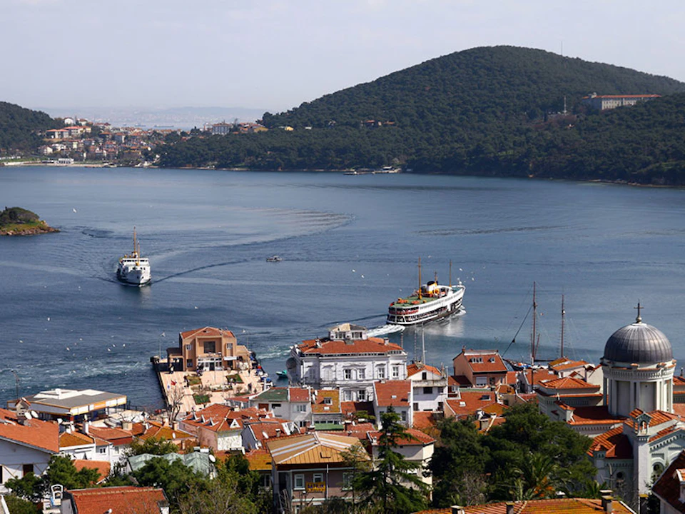 A coastal town with red-roofed buildings, a pier, and a ferry. Hills and a body of water with another ferry are in the background.