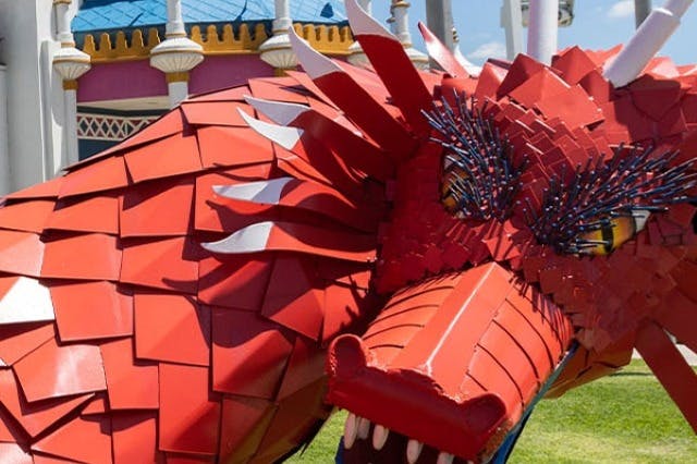 Detailní záběr sochy červeného draka s detailními šupinami a divokýma očima proti barevné budově s kopulemi a sloupy.