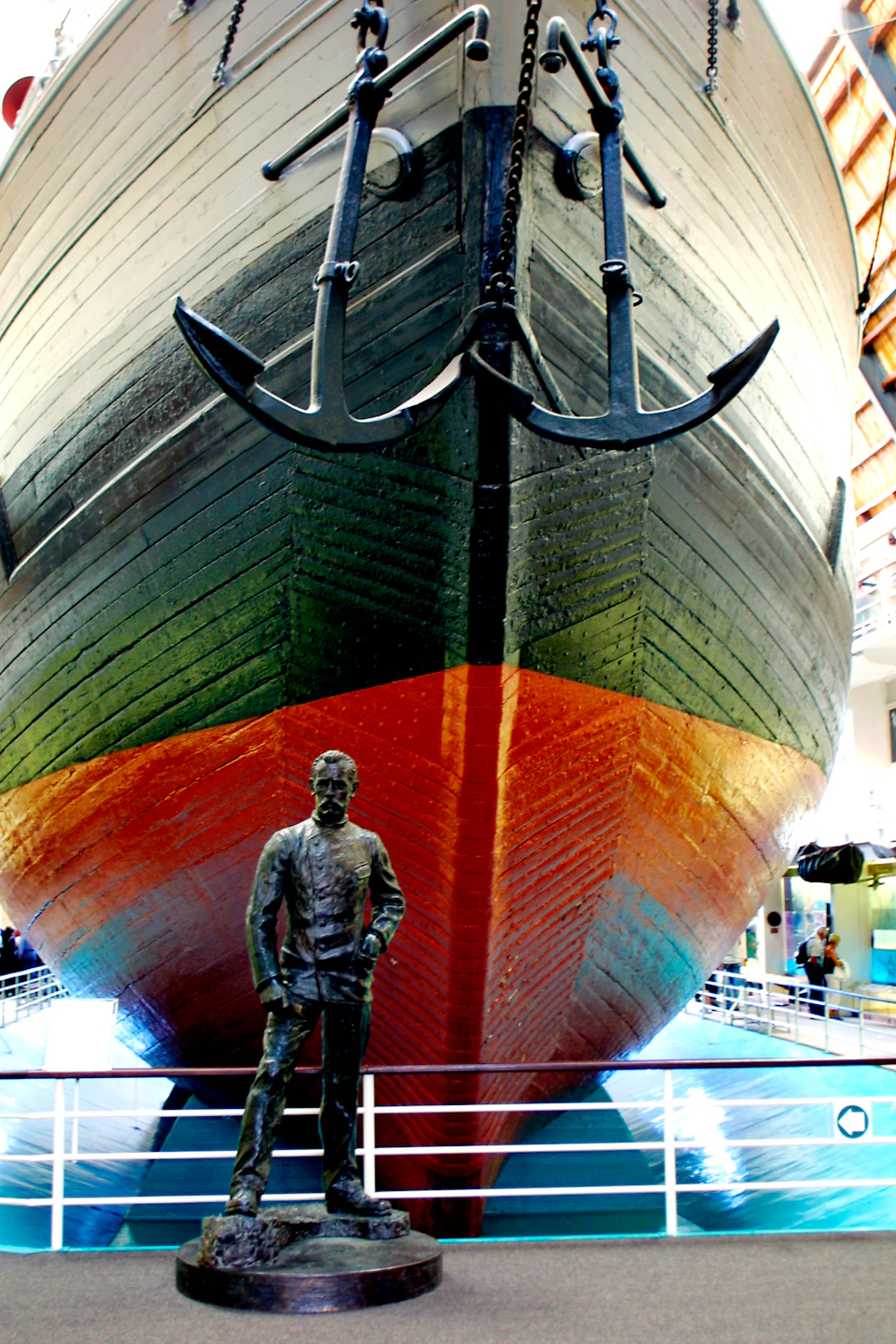 Large ship bow with dual anchors, displayed indoors above railings. Statue of a man in naval attire stands in front.