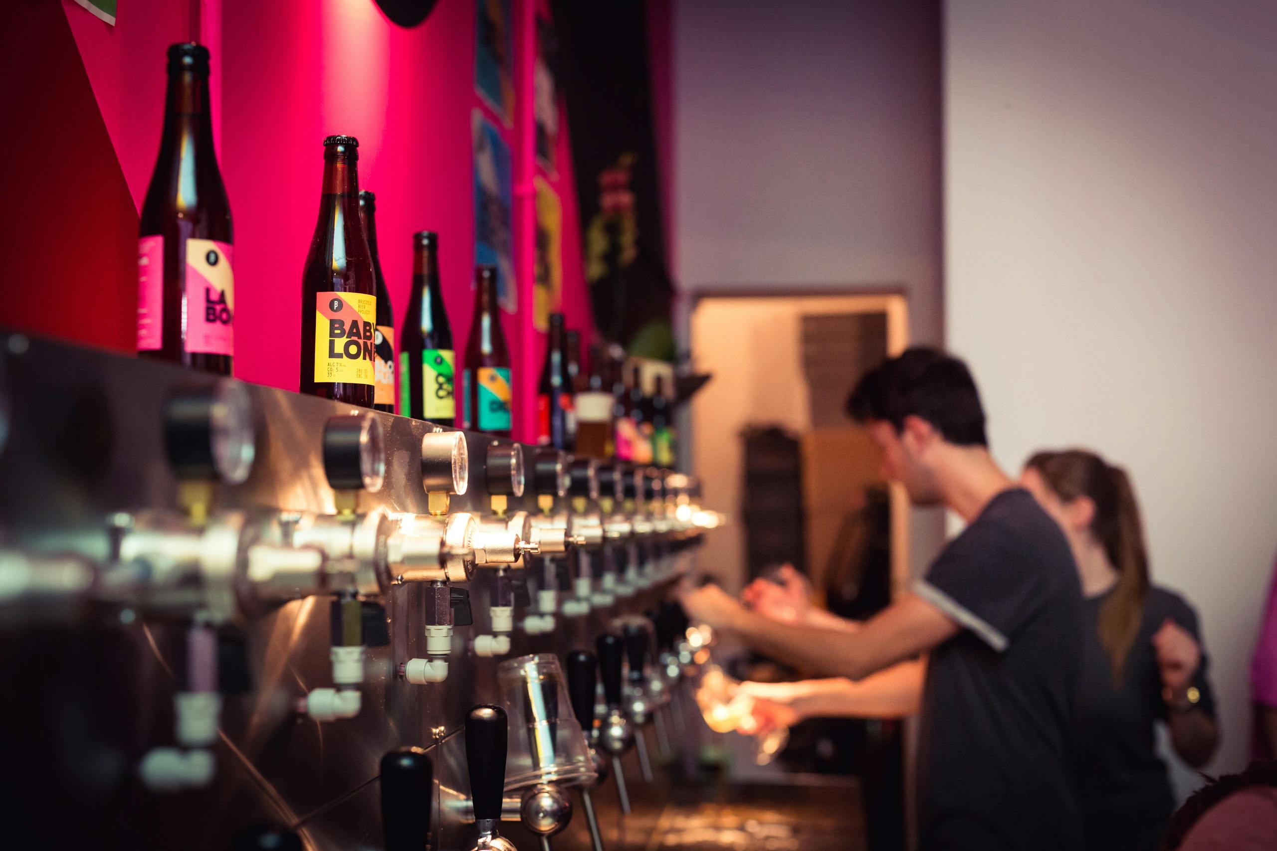 Un uomo versa un drink da una parete di rubinetti di birra con varie bottiglie colorate su uno scaffale in un bar poco illuminato.