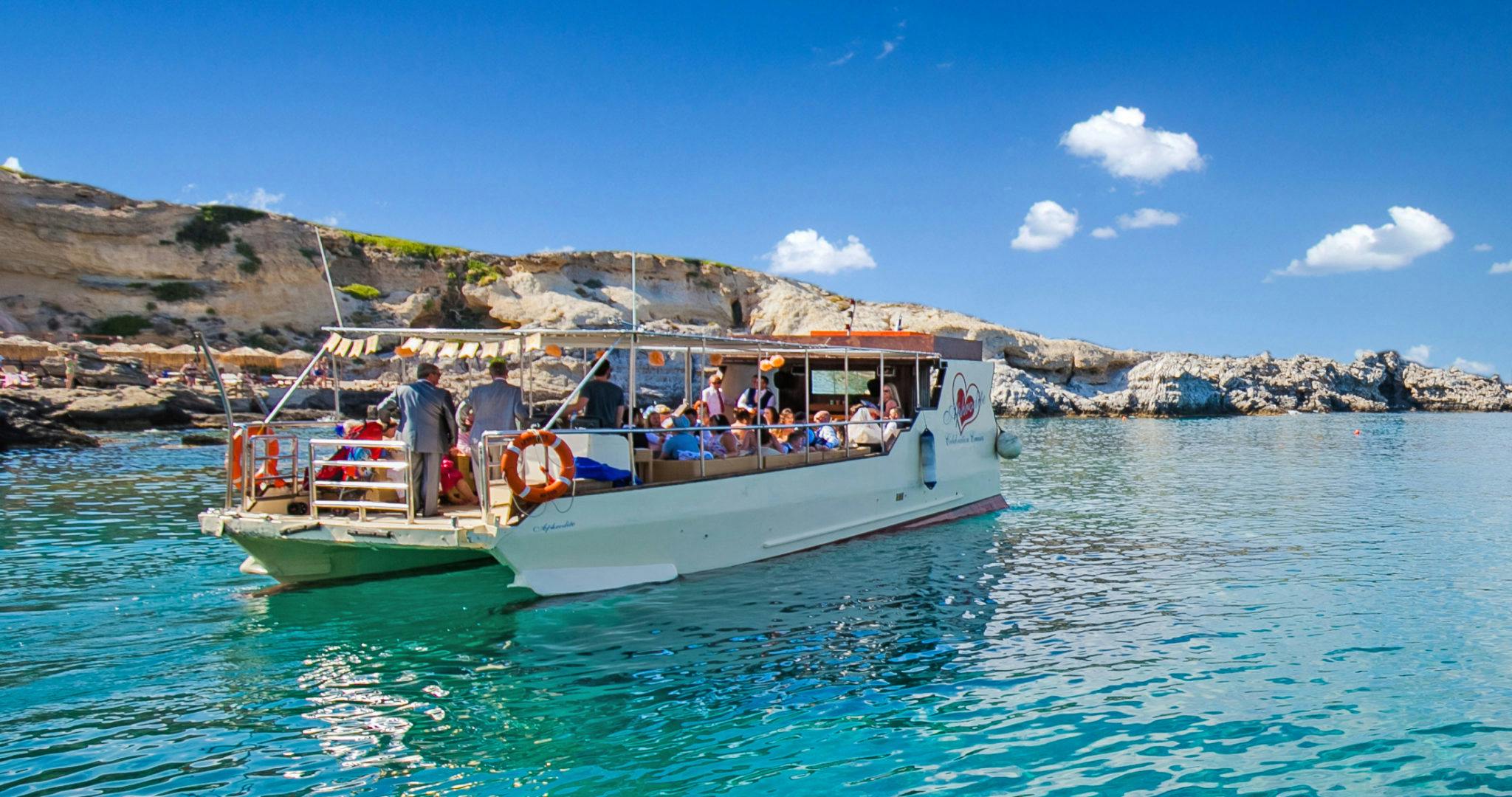 Una barca con passeggeri naviga su acque limpide vicino a scogliere rocciose sotto un cielo blu brillante.