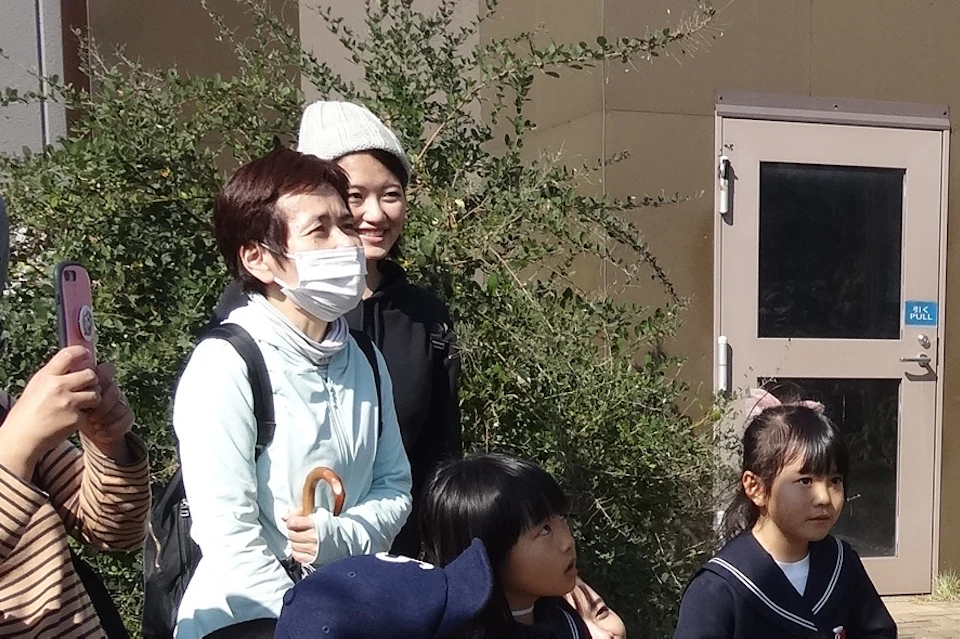 A group of people, including children in uniforms, observe and photograph a lemur climbing on an arm in an outdoor setting.