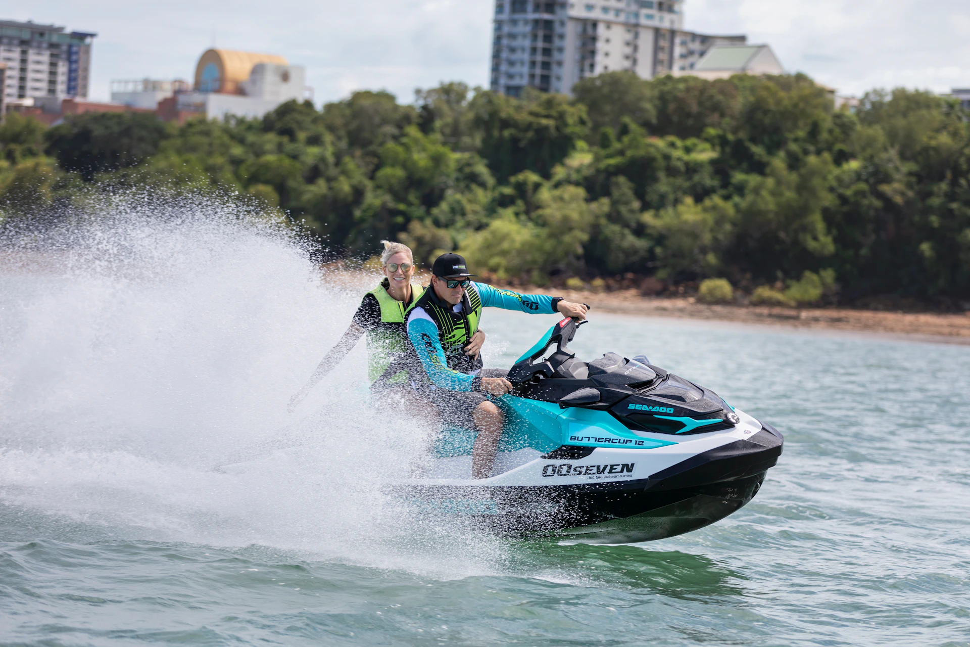 Two people ride a jet ski on water, both wearing life jackets. Trees and buildings are visible in the background.