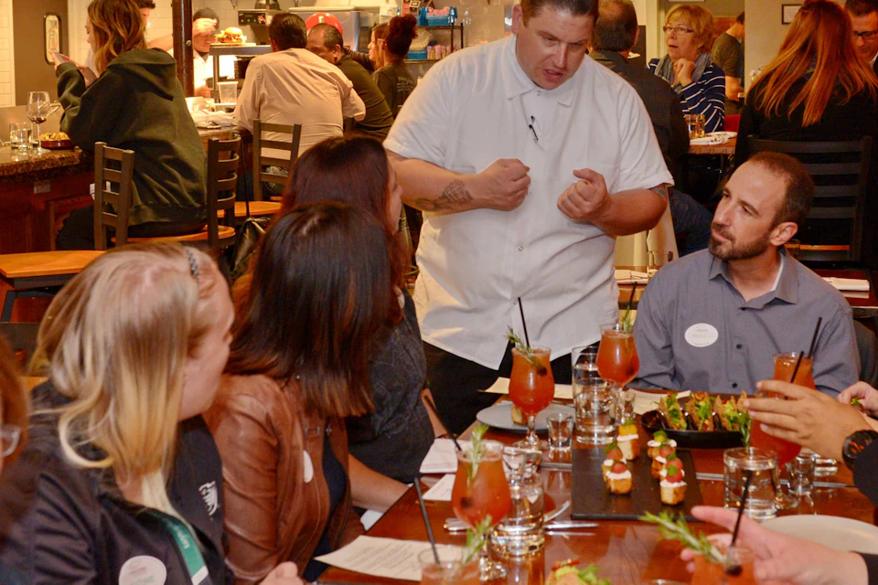 A group of people dining in a restaurant as a chef interacts with them, with food and drinks on the table.
