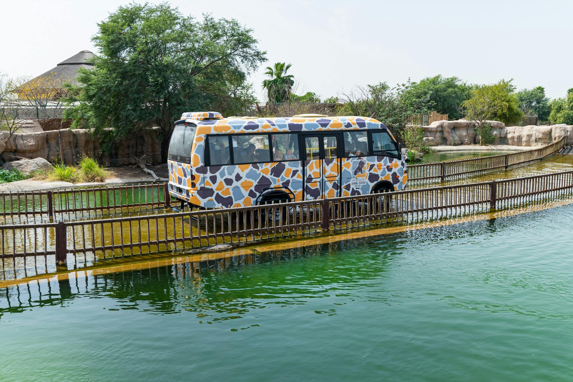 A bus with an animal-patterned exterior is crossing a bridge over water, with trees and greenery in the background.