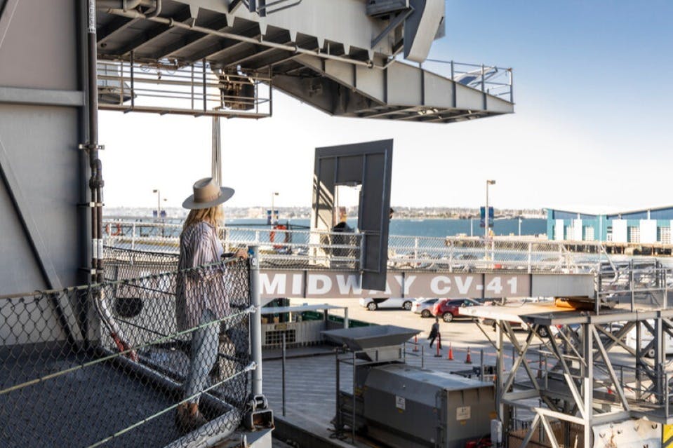 En person i hatt står på en utsiktsplattform ombord på USS Midway CV-41, med kustutsikt och hamn i bakgrunden.