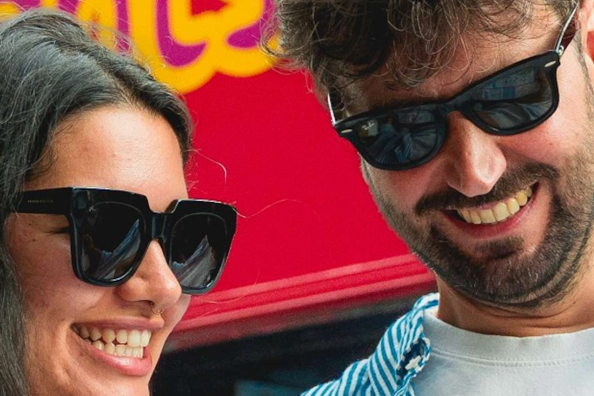 A man and a woman smiling while looking at a map in front of a red double-decker city sightseeing bus.