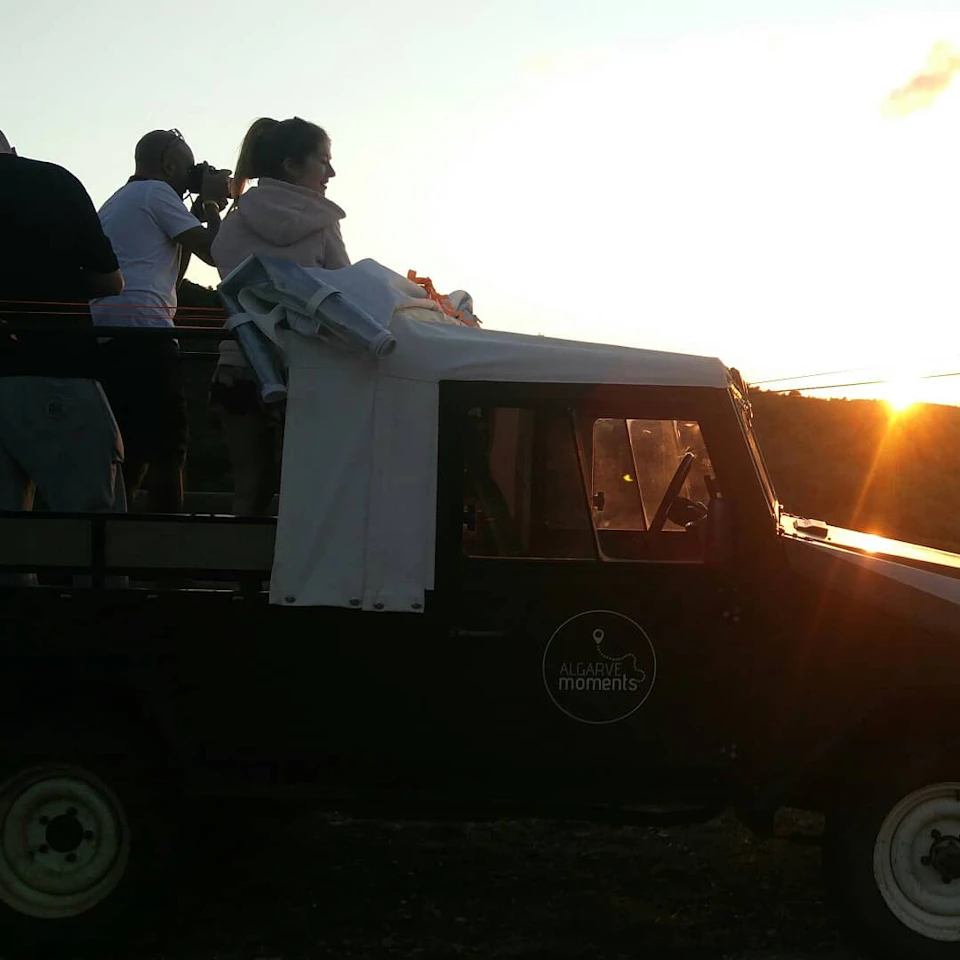 People stand in an open-top vehicle, one using binoculars, watching the sunrise or sunset in a natural setting.