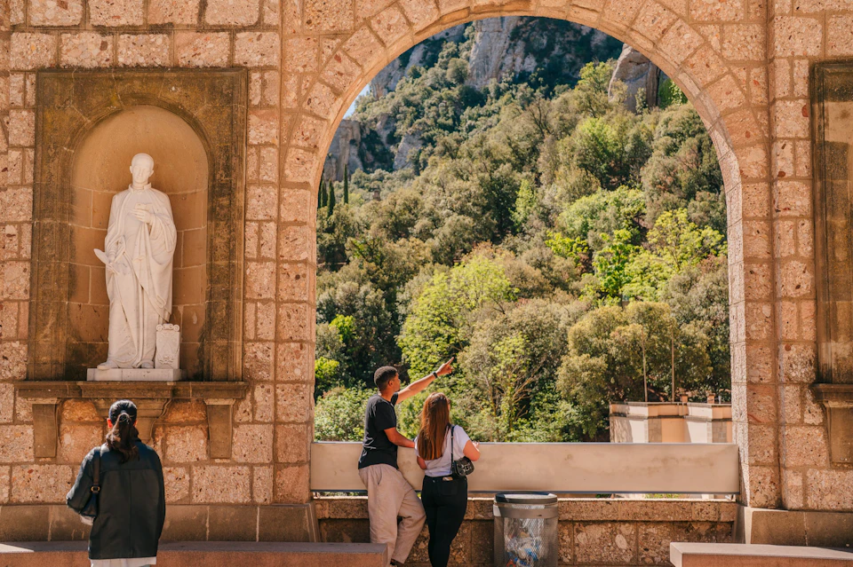 A man and a woman stand by a railing under a stone archway, overlooking a lush green forested hillside.