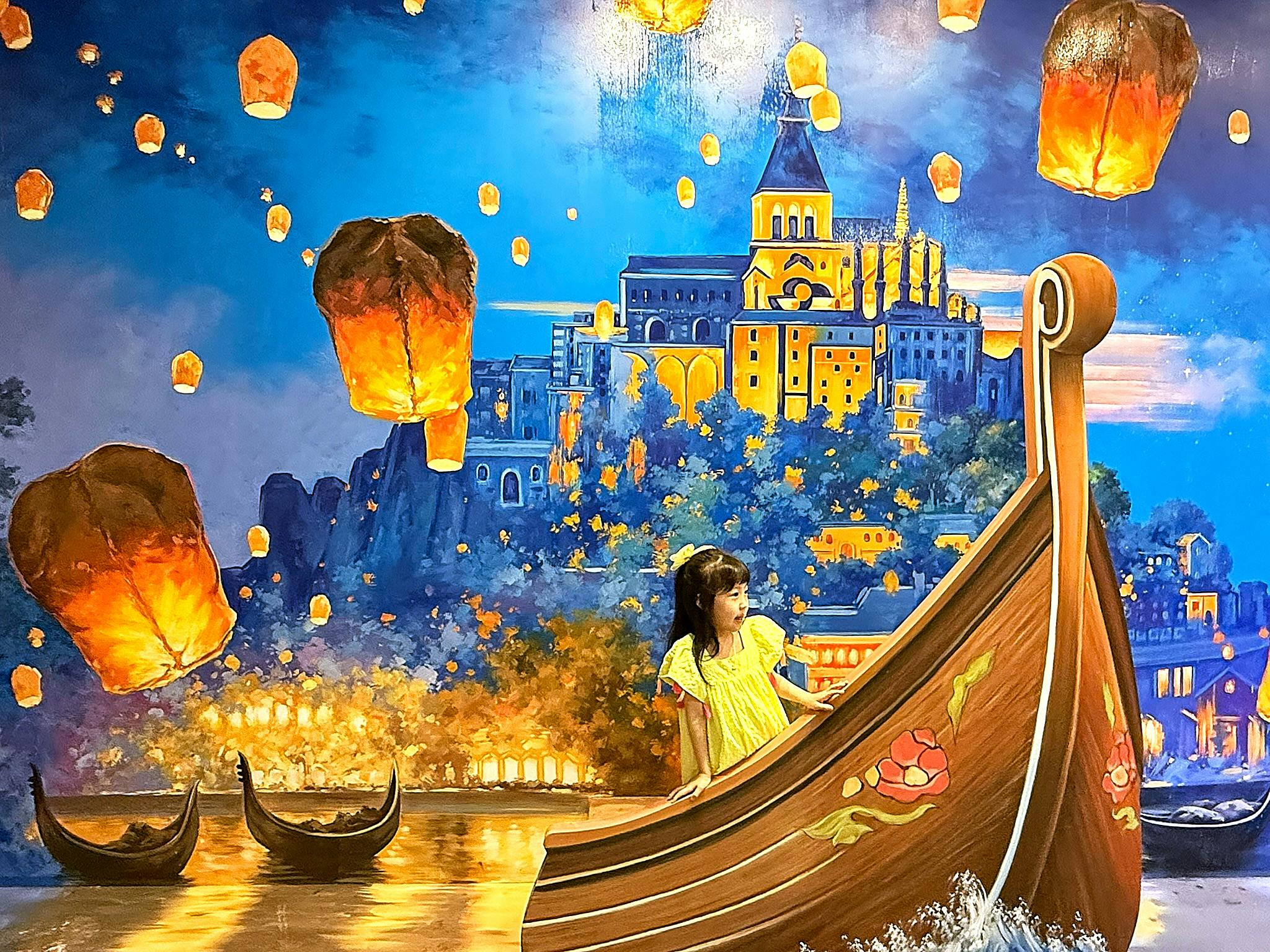 A girl in a yellow dress stands by a boat in front of a mural featuring floating lanterns and a castle at night.