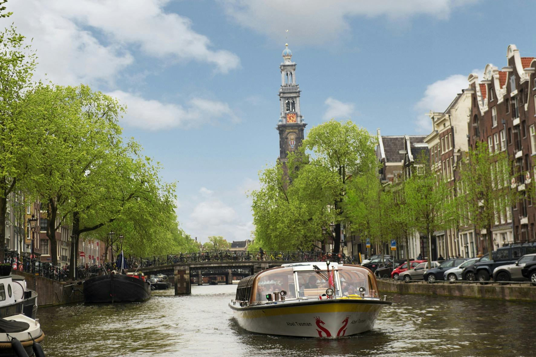 A canal boat navigates a waterway flanked by trees and historic buildings, with a clock tower visible in the background under a blue sky.