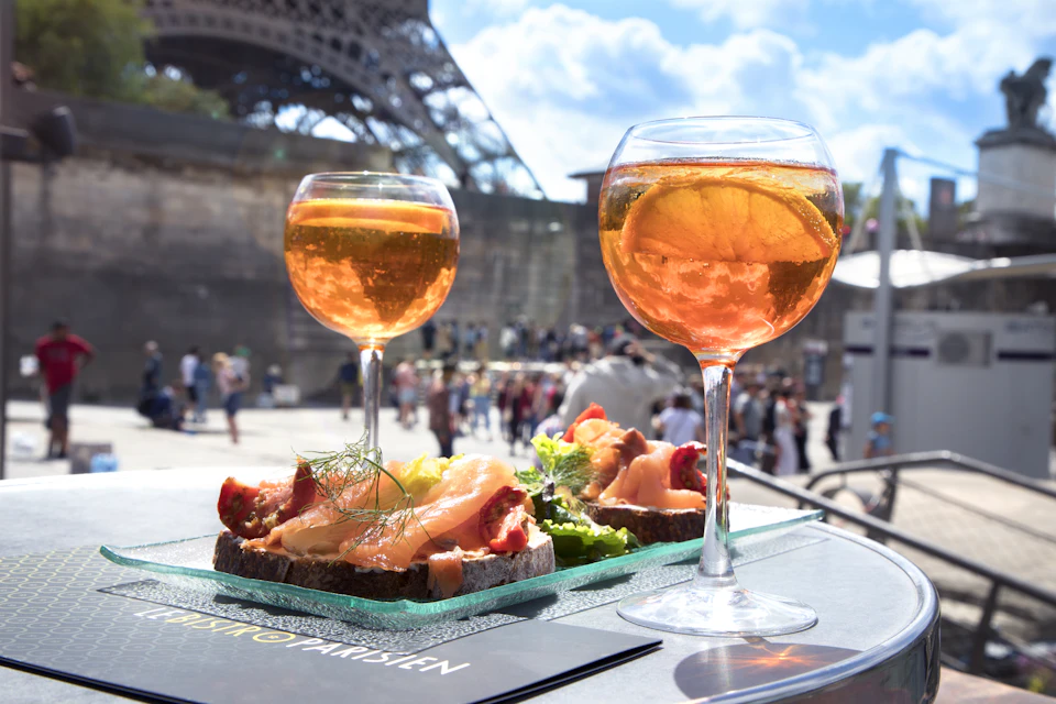 Two glasses of orange cocktails and plates of smoked salmon on toast with fresh garnish, with the Eiffel Tower in the background.